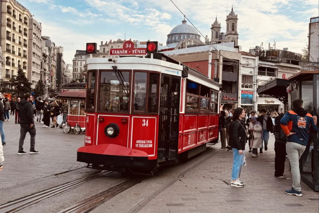 A classic, bright red vintage tram of Istanbul traveling down a pedestrian avenue lined with shops and historic buildings.
