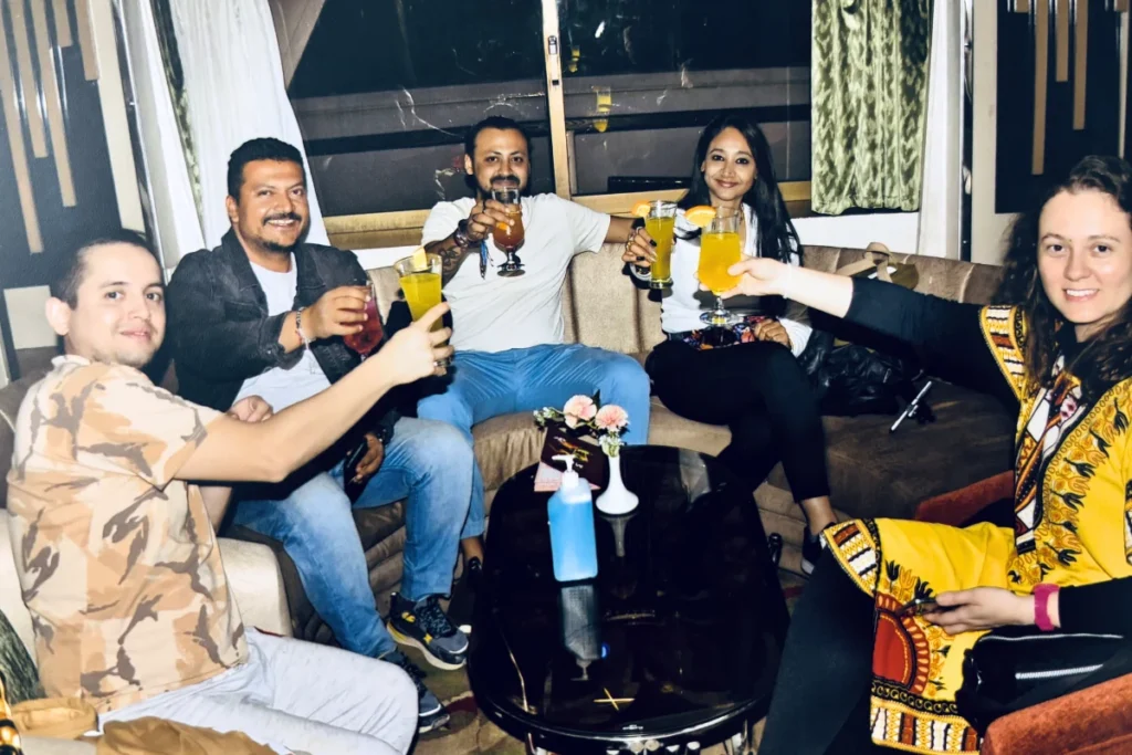 A group of five people sitting around a coffee table indoors, smiling and raising their glasses together in a joyful toast.