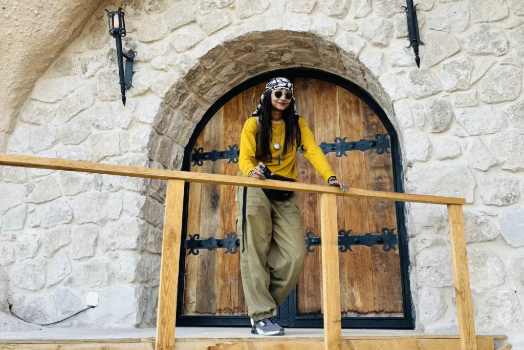 Sumana wearing a patterned scarf around their neck stands within a classic arched wooden doorway set into a textured stone wall.