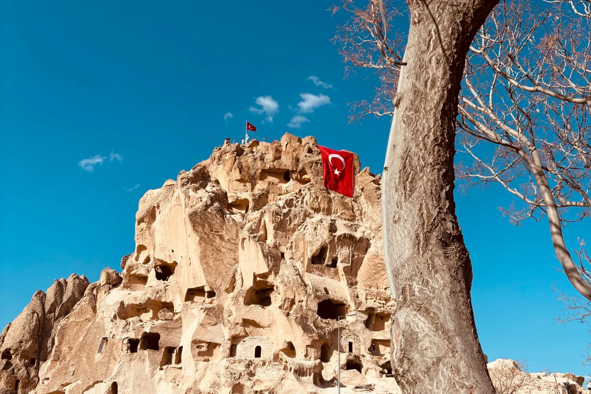 Panoramic view of the towering Uçhisar Castle rock fortress, featuring ancient carved cave openings and a Turkish flag mounted at its highest peak under a clear blue sky.