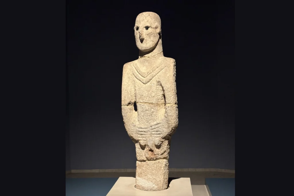 The ancient limestone "Urfa Man" statue, depicted with black stone eyes and hands clasped at the waist, displayed in a dark museum setting.