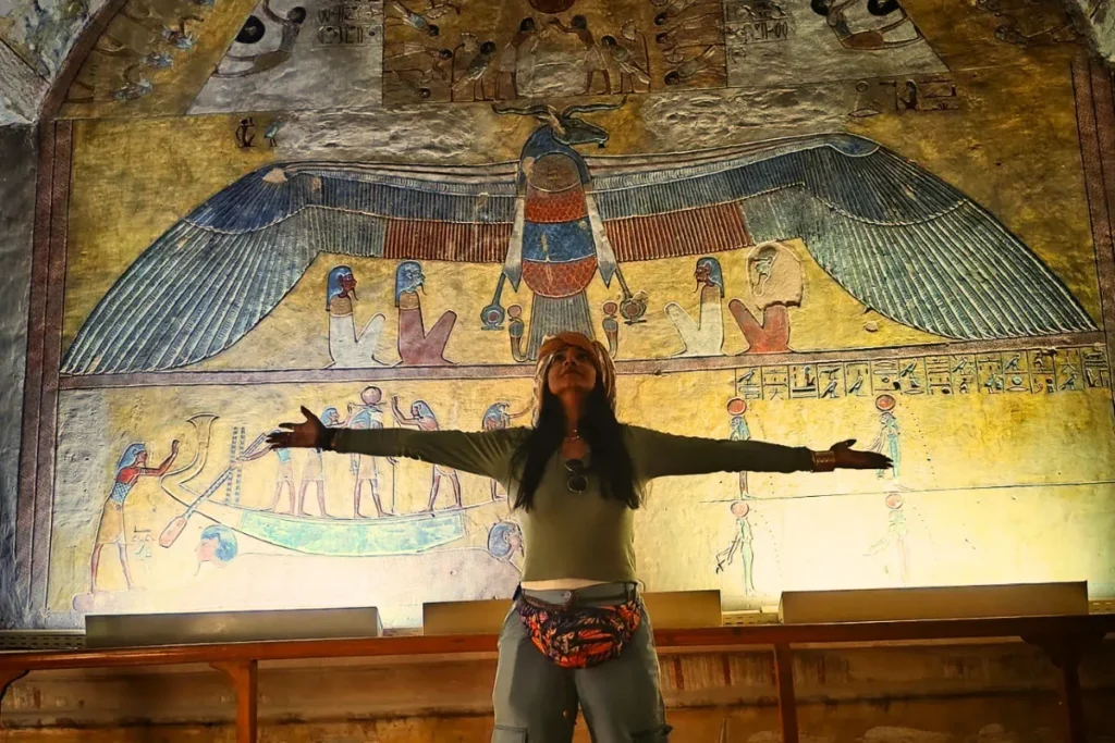 Sumana stands with arms wide open in the center of an underground Egyptian tomb room, facing vibrant and colorful hieroglyphic paintings spanning the historic walls.