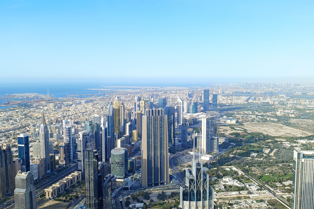 A wide aerial view of the sprawling Dubai skyline, featuring the dense cluster of high-rise skyscrapers along the coast next to the blue ocean.