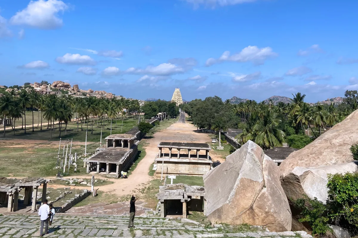 A wide panoramic view overlooking the ruined stone structures of the ancient bazaar market path leading towards the tall Virupaksha Temple gopuram tower in Hampi.