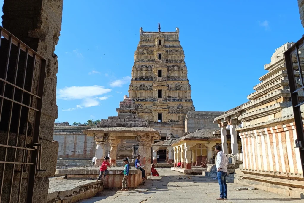 A daytime shot from the back of the Virupaksha Temple, showcasing its intricate multi-tiered stone tower against a blue sky.