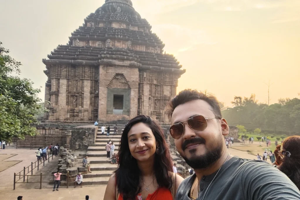 A selfie of Akash & Sumana smiling together with a historic Konark sun temple in the background.