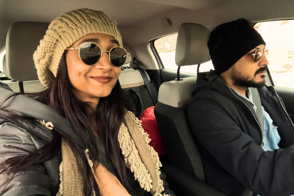 A close-up selfie of Akash & Sumana smiling inside a vehicle while wearing winter hats and sunglasses.