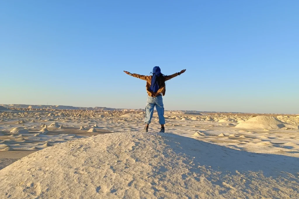 A high-angle shot of Sumana with outstretched arms leaping joyfully from a smooth white chalk mound under a clear blue sky.