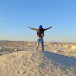 Sumana with her outstretched arms stands atop a smooth, white limestone hill in Egypt's White Desert under a bright sky.