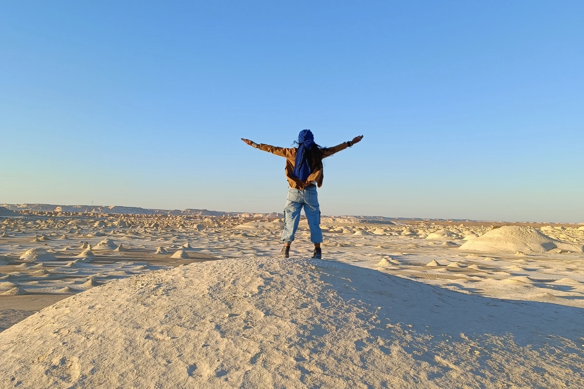 Sumana with her outstretched arms stands atop a smooth, white limestone hill in Egypt's White Desert under a bright sky.