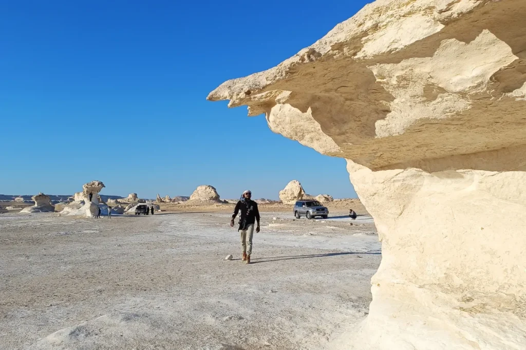 Akash walking past a striking, overhanging white chalk rock formation in a desolate desert landscape.