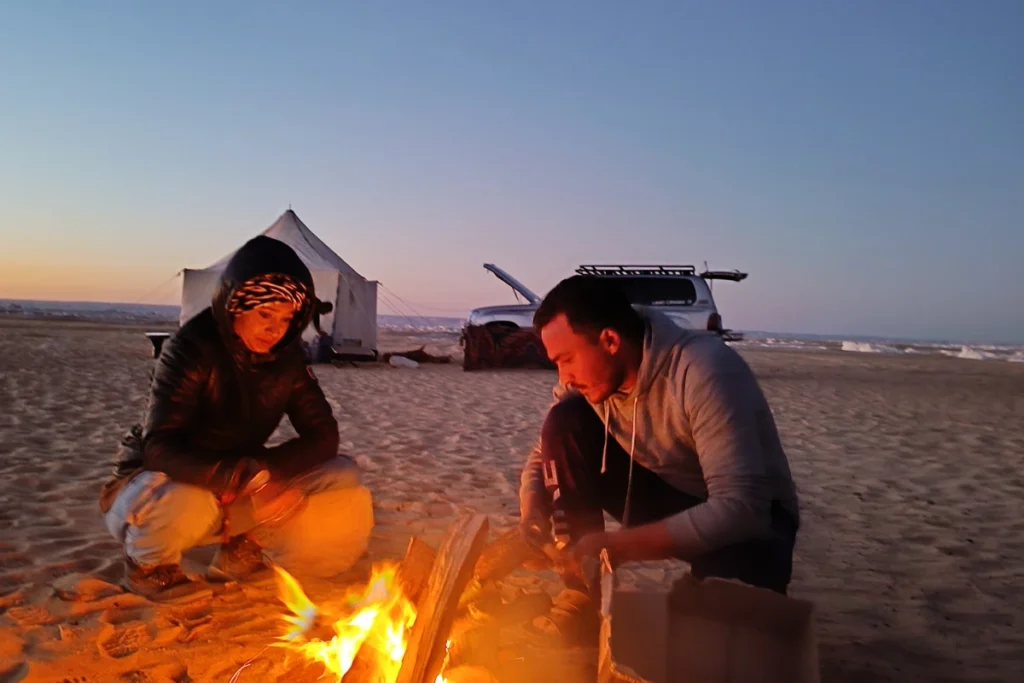 A night shot of a small desert campsite, showing Sumana and a local Bedouin sitting near a warm campfire next to a tent under a twilight sky.