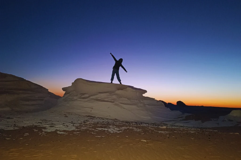 Silhouette of Sumana standing on a high white rock formation against a deep blue, pre-dawn horizon.