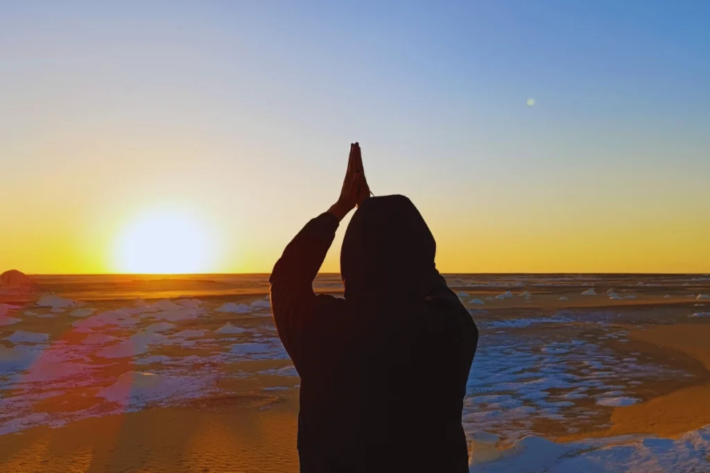 Silhouette of Akash with hands together in a peaceful pose, framed against a glowing orange and blue sunrise over the desert.