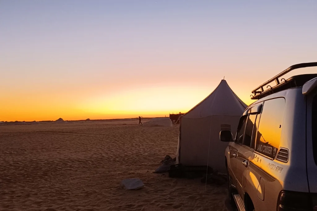 A camping setup in the white desert at dusk, featuring a canvas tent pitched next to an off-road vehicle.