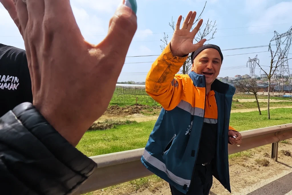 First-person perspective showing a high-five with a friendly Turkish highway worker wearing a bright yellow and orange high-visibility safety jacket.