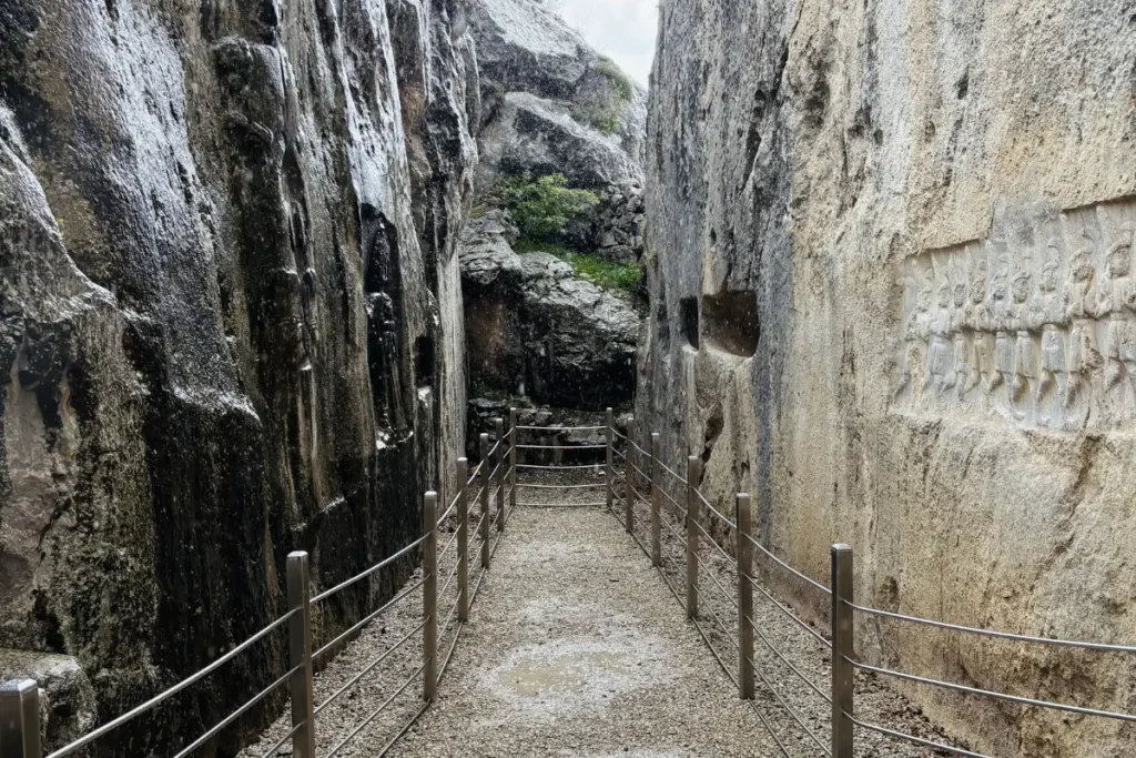A view looking down a narrow wooden walkway built between two sheer, towering rock faces adorned with reliefs.