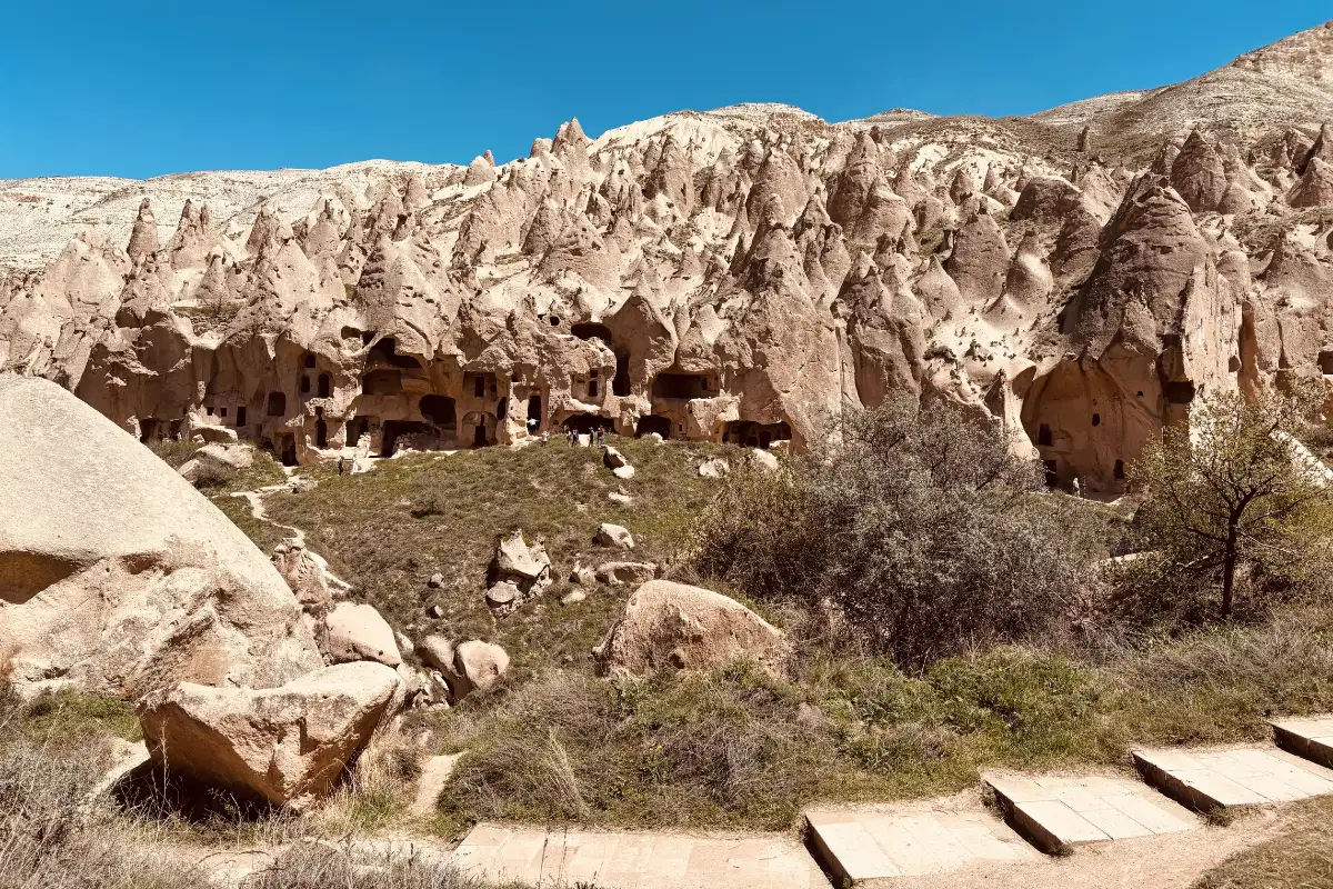 A wide landscape photograph showing a rugged cliffside filled with ancient hand-carved cave openings, rock dwellings, and natural stone formations surrounded by sparse green vegetation under bright daylight.