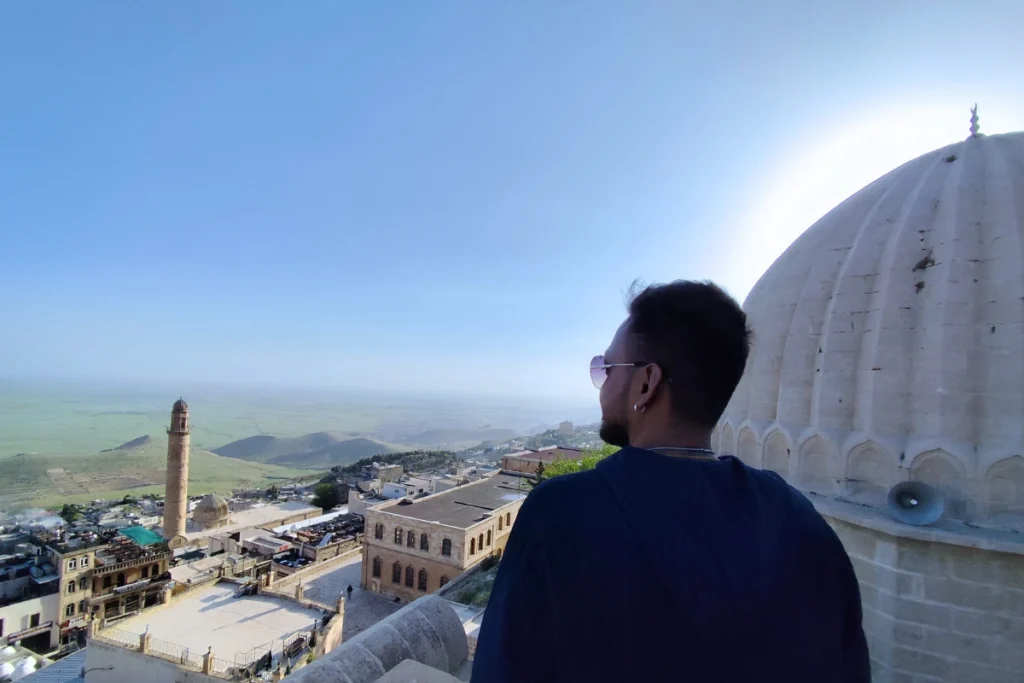  A back view of Akash with a blue shirt looking out over the historic city of Mardin under a clear blue sky.