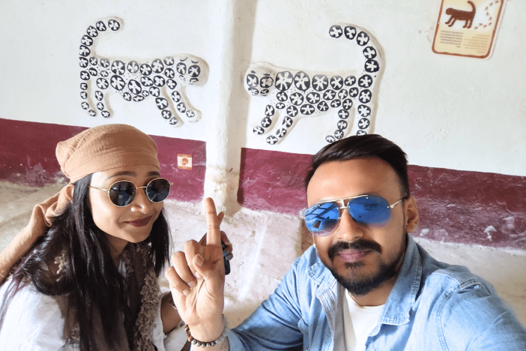 Akash & Sumana posing closely together in front of a white wall illustrated with ancient leopard artwork.