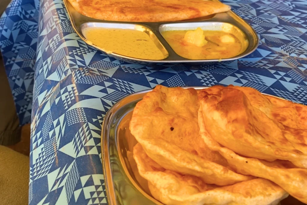 A stainless steel plate served with layered, golden-brown flaky puris next to a bowl of rich gravy at a roadside eatery.