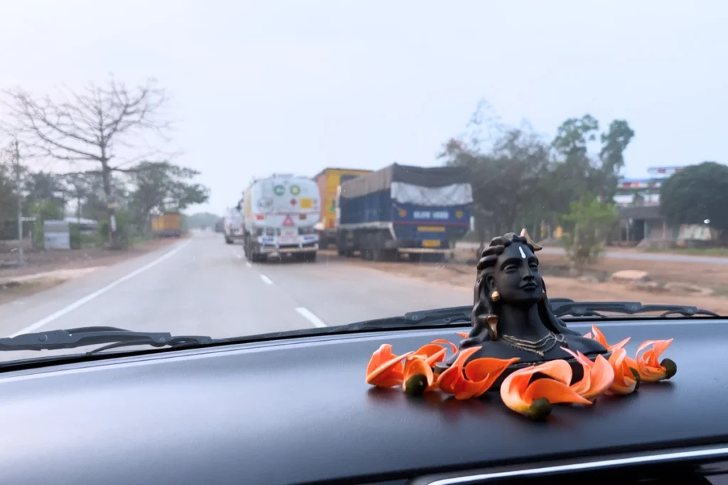 A shot from behind a motorcycle carrying cargo, driving on an open highway alongside other vehicles with a small shiva statue on the car dasboard.