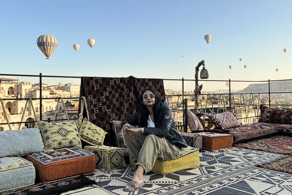 Sumana wearing sunglasses, a hoodie, and a dark jacket sits cross-legged on a yellow cushion on an outdoor rooftop terrace, posing for the camera with hot air balloons in the distant background.