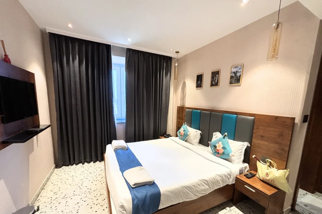 A modern hotel room at The Hive Hotel in Vizag, featuring a neatly made double bed with white linens, a blue bed runner, and matching blue-accented pillows. The room has a wooden headboard, a small bedside nightstand, dark floor-to-ceiling curtains, and a wall-mounted television across from the bed.