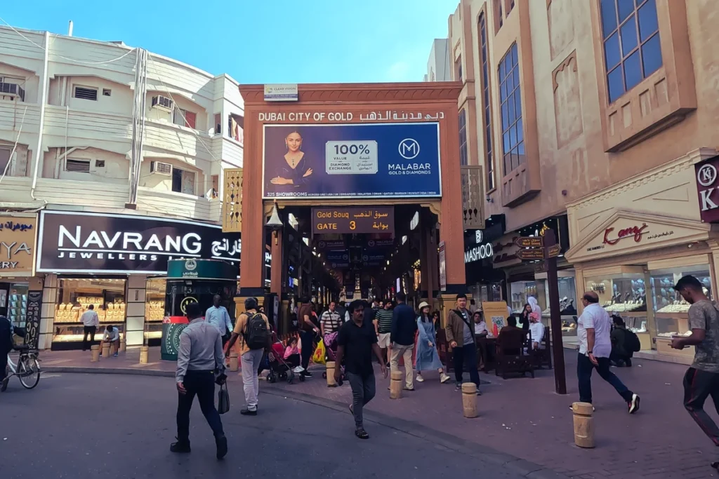 Dubai- Gold souk A daytime street-level shot of a busy market street with pedestrians walking past shops, highlighting a prominent sign for "Navrang Jewellers" at Dubai Gold Souk.