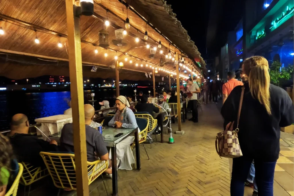 Dubai- creek restaurants 2 A side-angle view of people sitting on the benches at an outdoor waterfront area, looking out toward a pedestrians walking by.