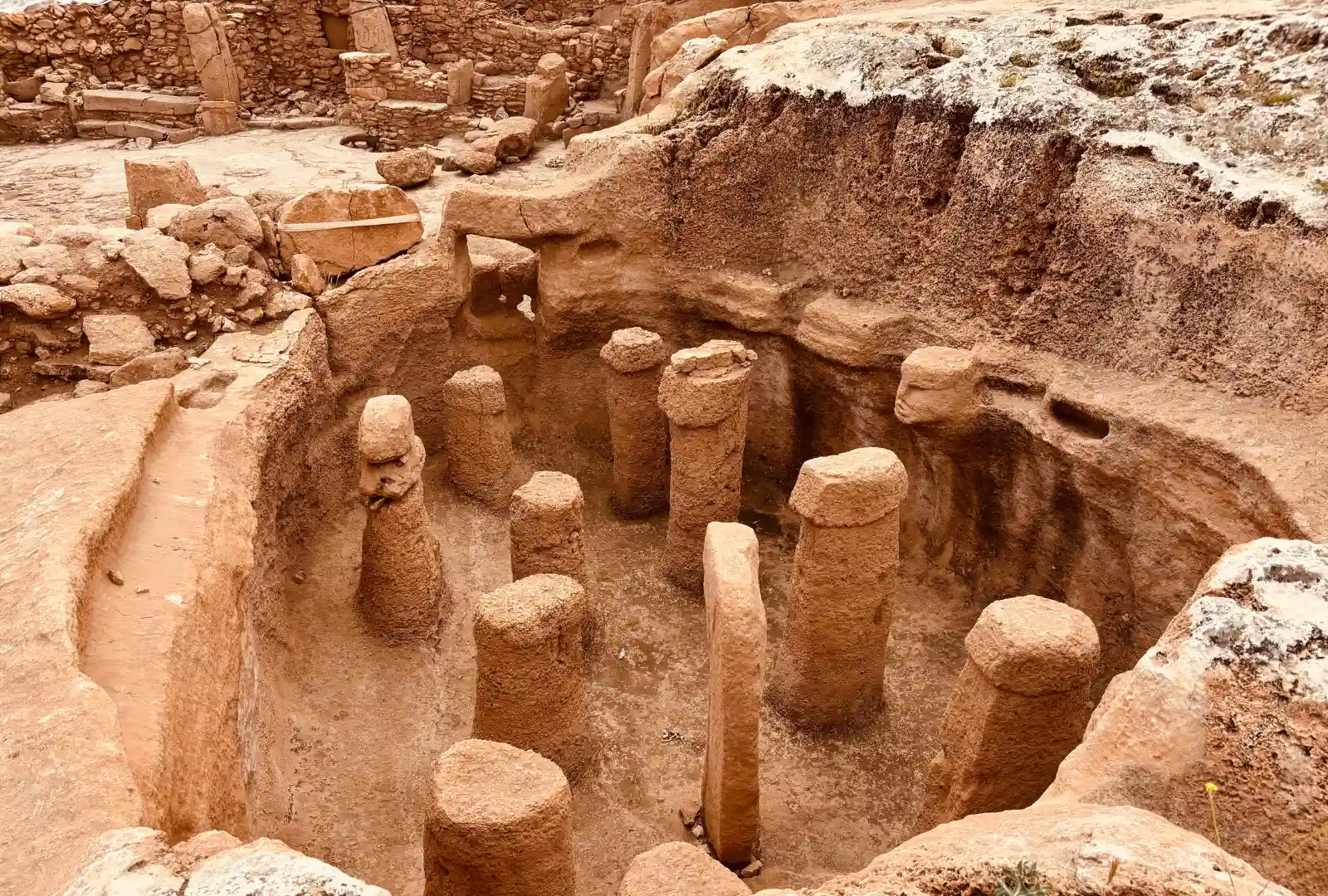 An overhead shot looking down into an excavated pit filled with ancient, carved stone pillars and a human face sculpture emerging from the bedrock.