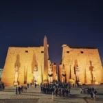 wide panoramic shot of the grand stone entrance to Luxor temple at night, heavily illuminated by spotlights, featuring massive pylon walls, monumental statues standing guard, and a tall central obelisk against a deep blue evening sky.