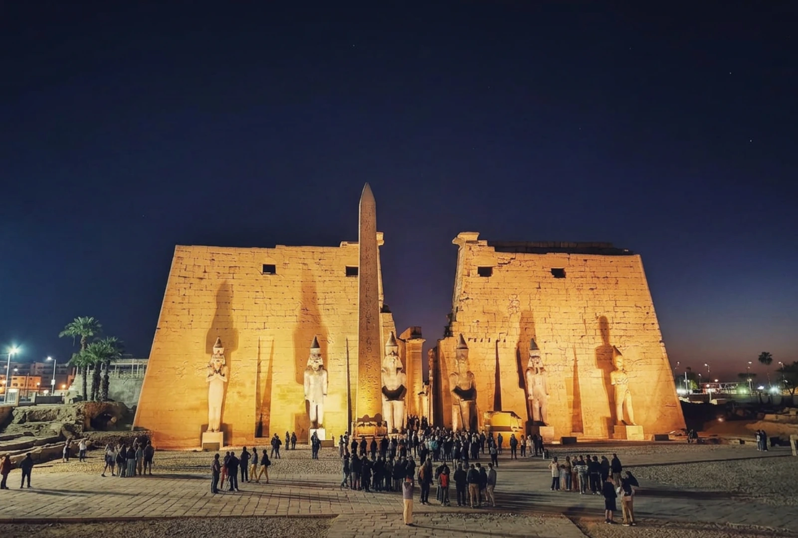 wide panoramic shot of the grand stone entrance to Luxor temple at night, heavily illuminated by spotlights, featuring massive pylon walls, monumental statues standing guard, and a tall central obelisk against a deep blue evening sky.