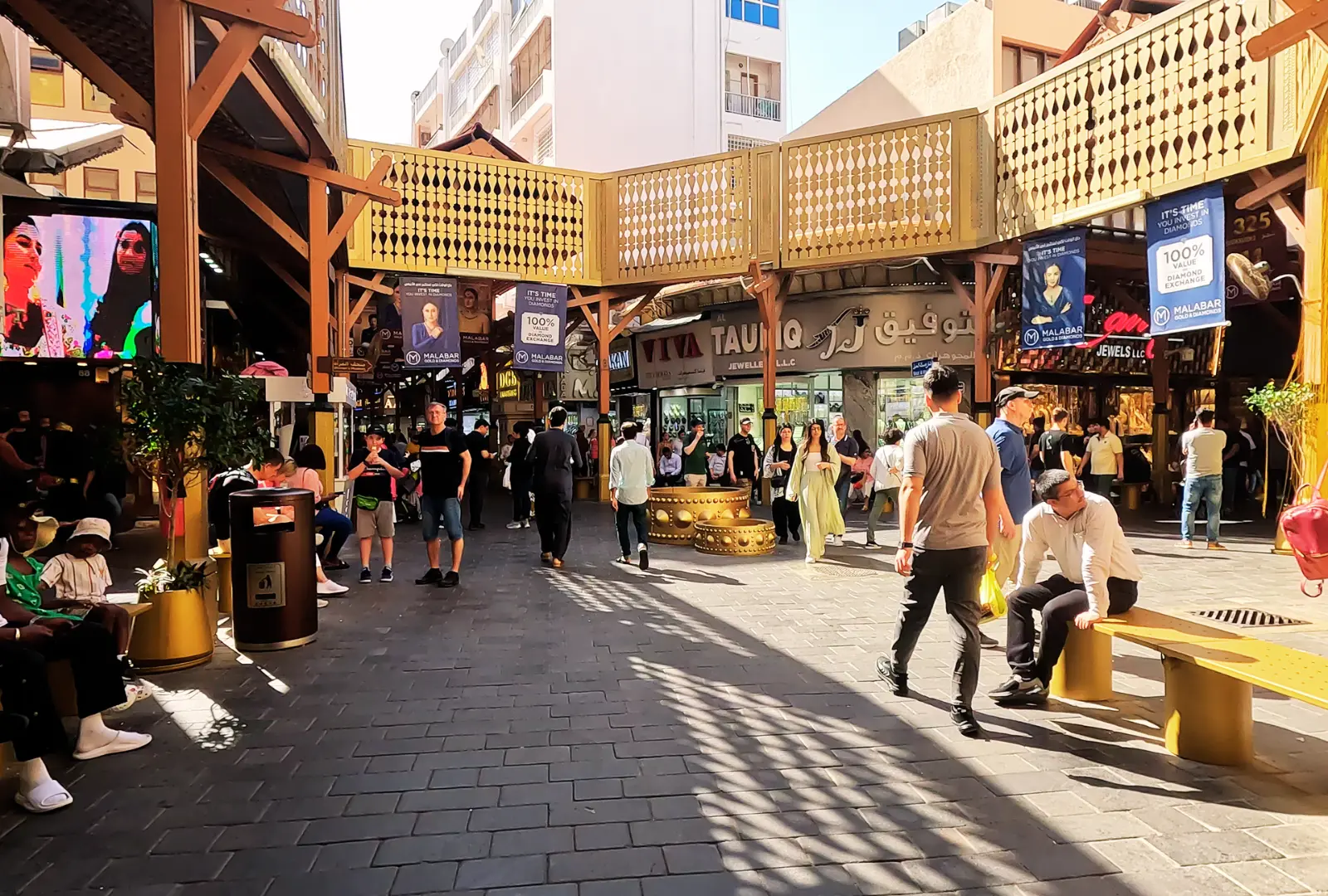 A wide landscape shot showing a crowded Gold Souk with people walking, sitting on benches, and shopping outside various storefronts in Dubai.