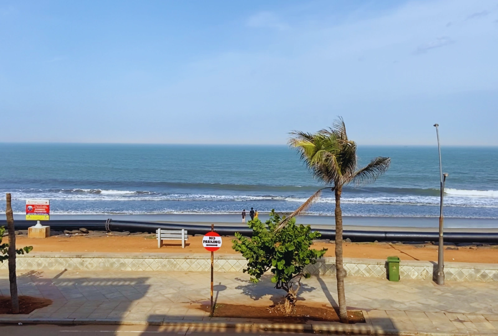 A wide view of a sandy beach meeting the ocean waves under a blue sky, featuring a palm tree on the right and a "No Parking" sign in the foreground.