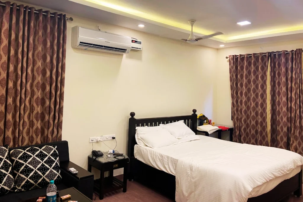 Pondicherry- Pranav Hotel room A neat bedroom featuring a large bed, air conditioner, and white walls with a dark wooden door.