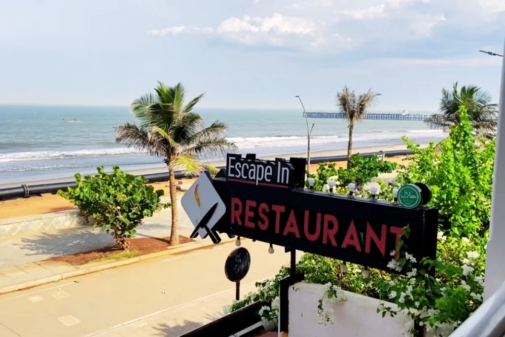 Pondicherry- View from Escape In Looking through a leafy archway at a restaurant sign near the sandy beach area.
