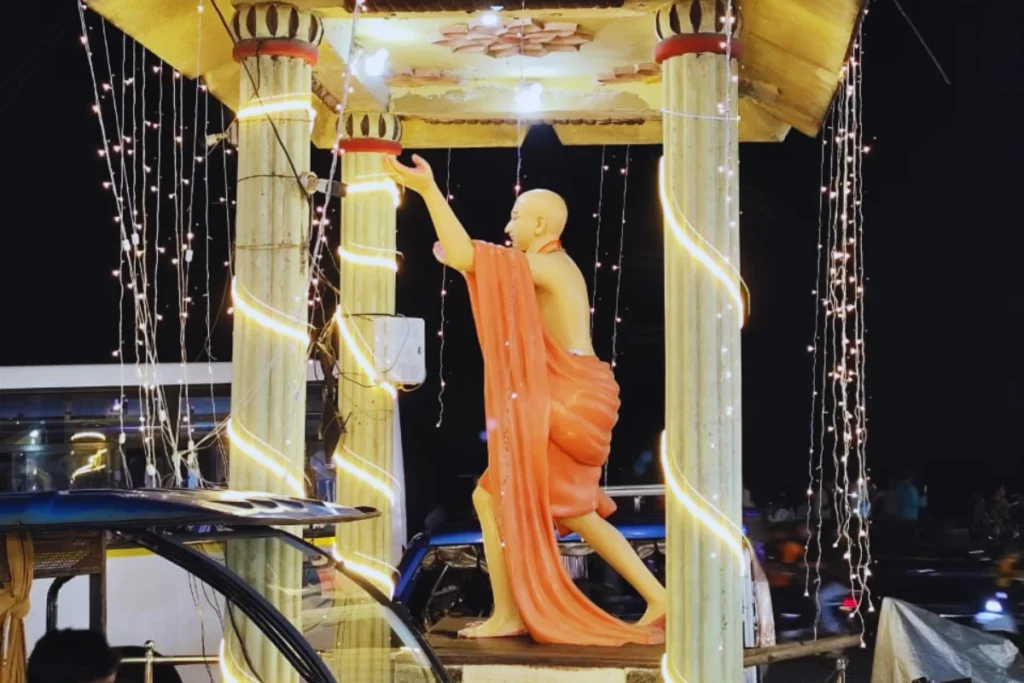 Puri- Chaitanya Statute A tall, bright orange statue of Saint Chaitanya standing on a pedestal adorned with string lights at night.
