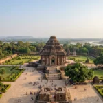 A high-angle wide landscape shot showing the grand stone structure of the Konark Sun Temple surrounded by green lawns and trees under a cloudy sky.