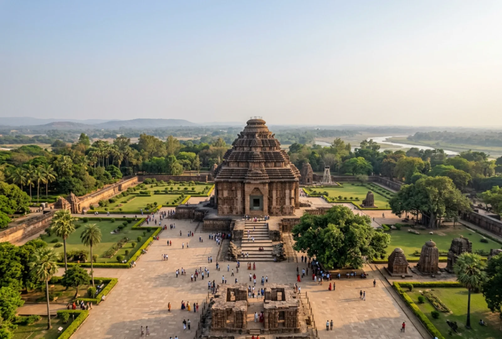 A high-angle wide landscape shot showing the grand stone structure of the Konark Sun Temple surrounded by green lawns and trees under a cloudy sky.
