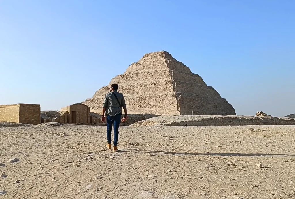 SAQQARA-1 Akash walks across the desert sand toward the grand facade of the Step Pyramid in the background.