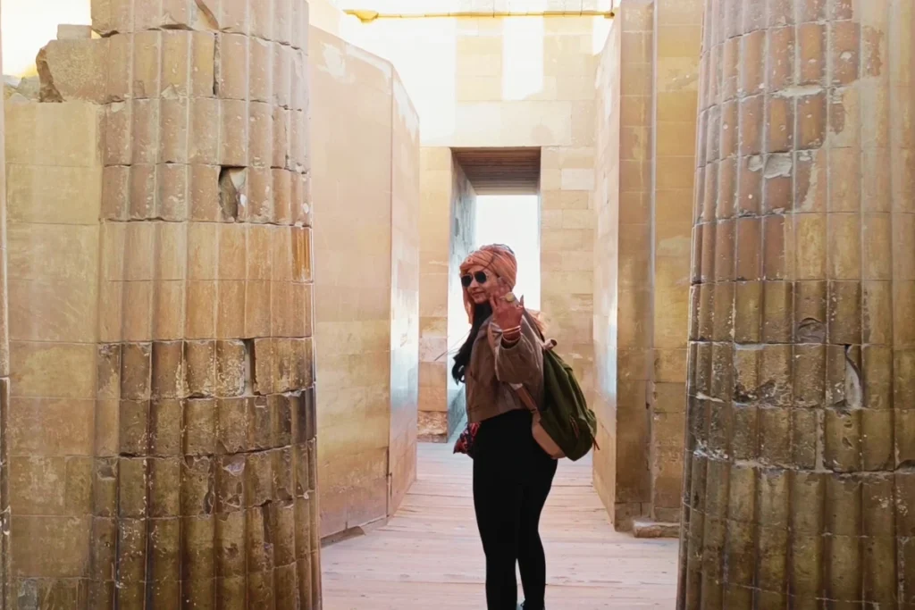 SAQQARA- 4 Sumana in a headscarf stands between two rows of massive, ancient stone pillars inside the illuminated entrance hall of the Saqqara complex.