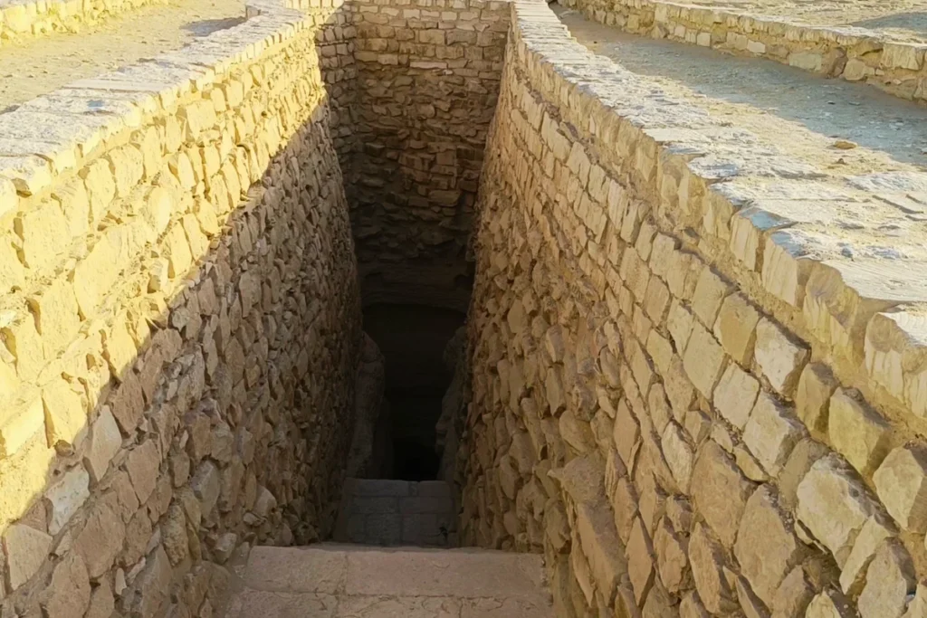 SAQQARA- 6 A square stone opening in the ground reveals a steep, stone-walled staircase leading down into an underground tunnel.