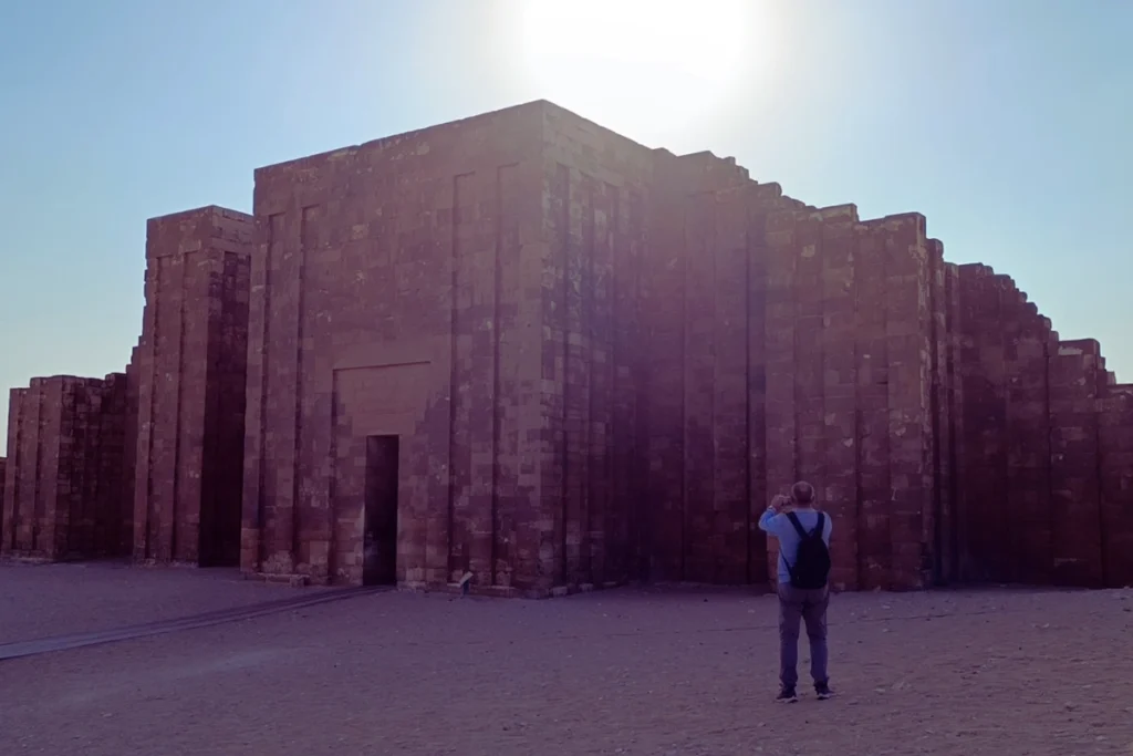 SAQQARA- 7 A high, block-like stone wall featuring a narrow, grand entrance gateway under a bright blue sky.