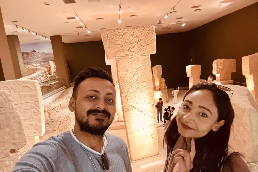A selfie of Akash & Sumana smiling together indoors in front of the large T-shaped stone pillars in the Göbekli Tepe replica room.
