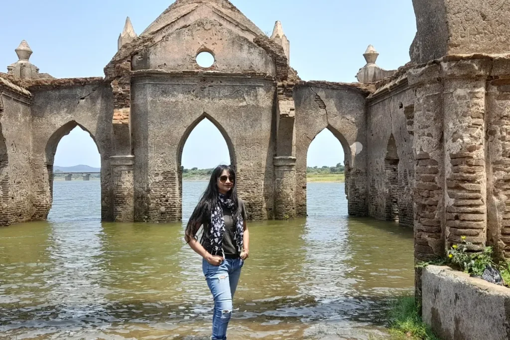 Settiheli Church-1 Sumana wearing sunglasses and a scarf poses in front of the flooded stone archways of the ruined Shettihalli Rosary Church.