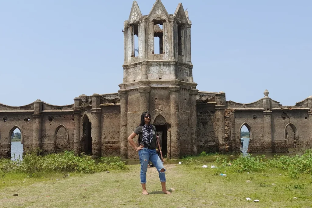 Settiheli Church-2 Sumana standing in front of The Gothic-style stone ruins of the Shettihalli Rosary Church standing partially submerged in water under a blue sky.