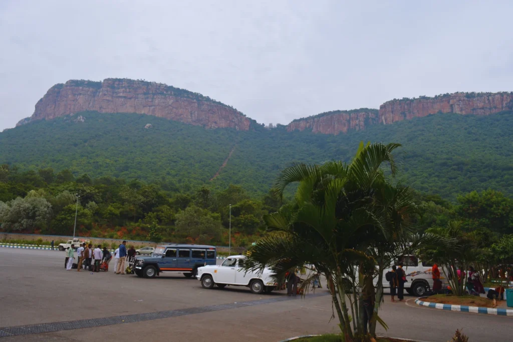 Tirupati- By bus stand A view from a parking lot or transit area looking toward a massive, green mountain peak under an overcast sky.