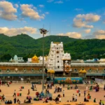 A wide horizontal landscape shot showing the grand white tiered tower of the Tirumala Venkateswara Temple complex situated against lush green hills under a cloudy sky.