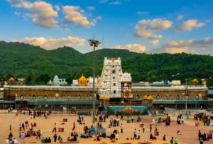 A wide horizontal landscape shot showing the grand white tiered tower of the Tirumala Venkateswara Temple complex situated against lush green hills under a cloudy sky.